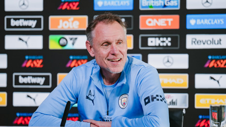 A Manchester City coach in a light blue tracksuit speaks at a press conference with a backdrop of sponsor logos.