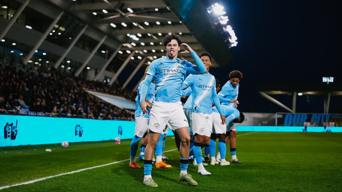 A group of football players in light blue jerseys celebrates a goal on a soccer field. The stadium is filled with spectators, and an LED screen displays a message. The atmosphere is lively under stadium lights.