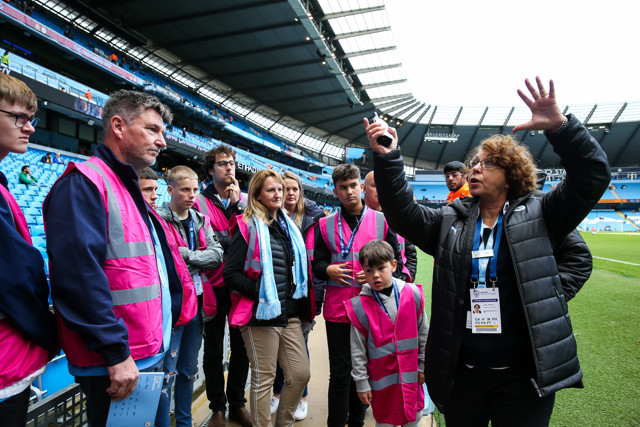 A tour guide is explaining to a group of people inside the Etihad Stadium, with most members of the group wearing pink vests.
