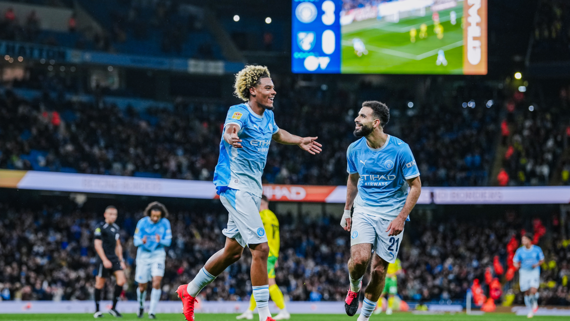 Two Manchester City players celebrating a goal during a match at Etihad Stadium with the scoreboard showing a 3-0 lead.