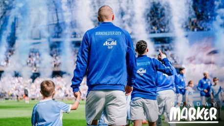 Manchester City players wearing blue jackets walk onto the field holding hands with young mascots. The background features smoke and a large crowd. The image has the text 'Monday Marker' at the bottom right.