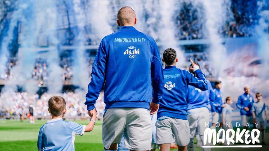 Manchester City players wearing blue jackets walk onto the field holding hands with young mascots. The background features smoke and a large crowd. The image has the text 'Monday Marker' at the bottom right.