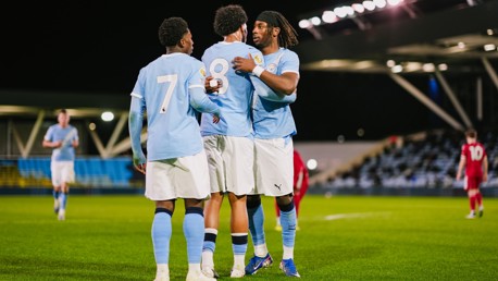 Three football players in light blue jerseys, numbers 7, 8, and another player, embrace on the field likely celebrating a goal. The stadium is lit at night with players in red jerseys in the background.
