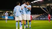 Three football players in light blue jerseys, numbers 7, 8, and another player, embrace on the field likely celebrating a goal. The stadium is lit at night with players in red jerseys in the background.