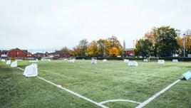 A football pitch with small portable goals set up, surrounded by residential houses and trees with autumn foliage.