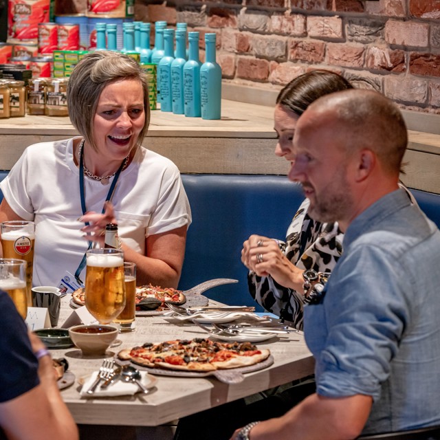 A group of people sitting around a table in a restaurant, sharing pizza and drinks. They appear to be having a lively conversation.