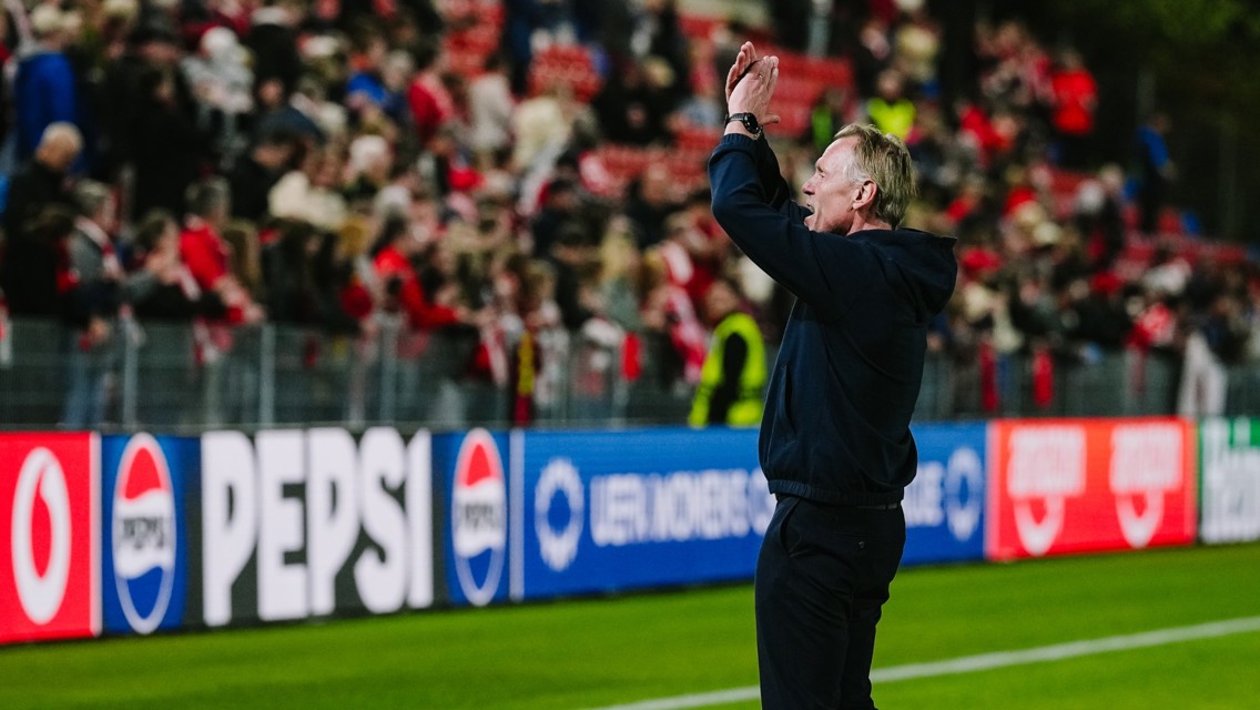 A soccer coach stands on the sidelines, applauding towards a crowd in a stadium. The crowd is dressed in red and there are Pepsi advertisements visible.