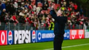 A soccer coach stands on the sidelines, applauding towards a crowd in a stadium. The crowd is dressed in red and there are Pepsi advertisements visible.