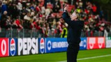 A soccer coach stands on the sidelines, applauding towards a crowd in a stadium. The crowd is dressed in red and there are Pepsi advertisements visible.