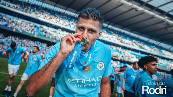 A football player in a blue Manchester City jersey kisses a Premier League medal on the pitch. The background shows a stadium full of spectators.