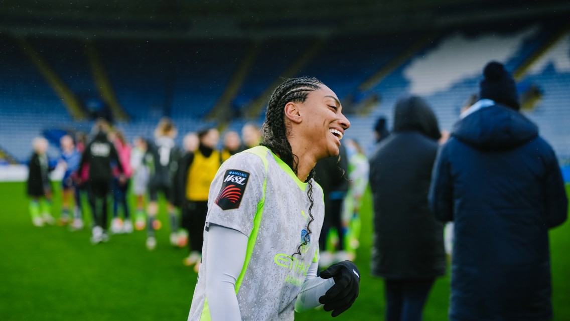 A Manchester City Women's Team player in a rain-spotted uniform with Barclays WSL patch on the sleeve walks off the pitch. Background shows other players and staff on the field.
