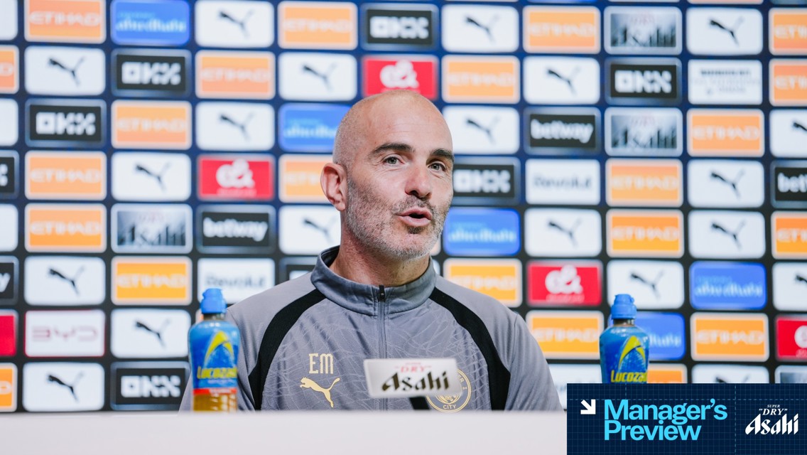 A person speaking at a press conference with a backdrop showcasing various sponsor logos such as Puma and Etihad. Two bottles of Lucozade are positioned on the table near the microphone labeled Asahi.