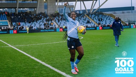 Football player wearing Manchester City Women's kit celebrates after scoring 100 goals, holding a colorful ball on a football field, with stadium seating and scoreboard showing Man City Women vs. Liverpool.