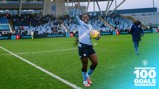 Football player wearing Manchester City Women's kit celebrates after scoring 100 goals, holding a colorful ball on a football field, with stadium seating and scoreboard showing Man City Women vs. Liverpool.