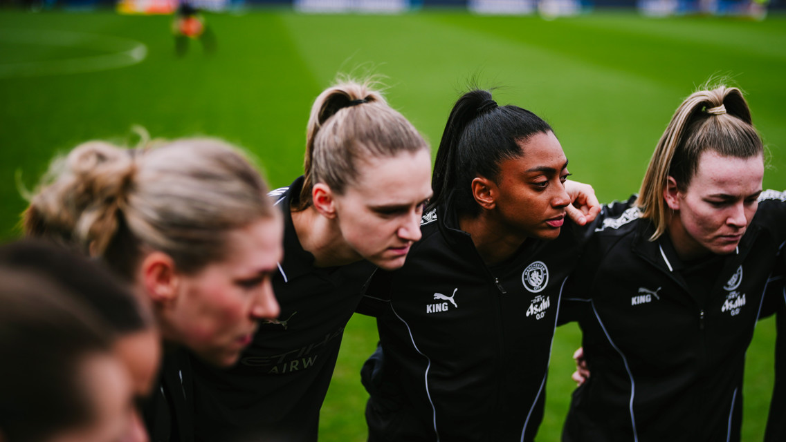 A women's football team in a huddle on a grass pitch wearing black PUMA tracksuits with Manchester City logo.
