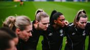 A women's football team in a huddle on a grass pitch wearing black PUMA tracksuits with Manchester City logo.