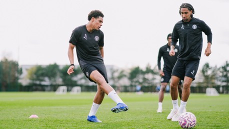 Three football players from Manchester City's youth team training on a grassy field, engaging in a dribbling exercise with a football.