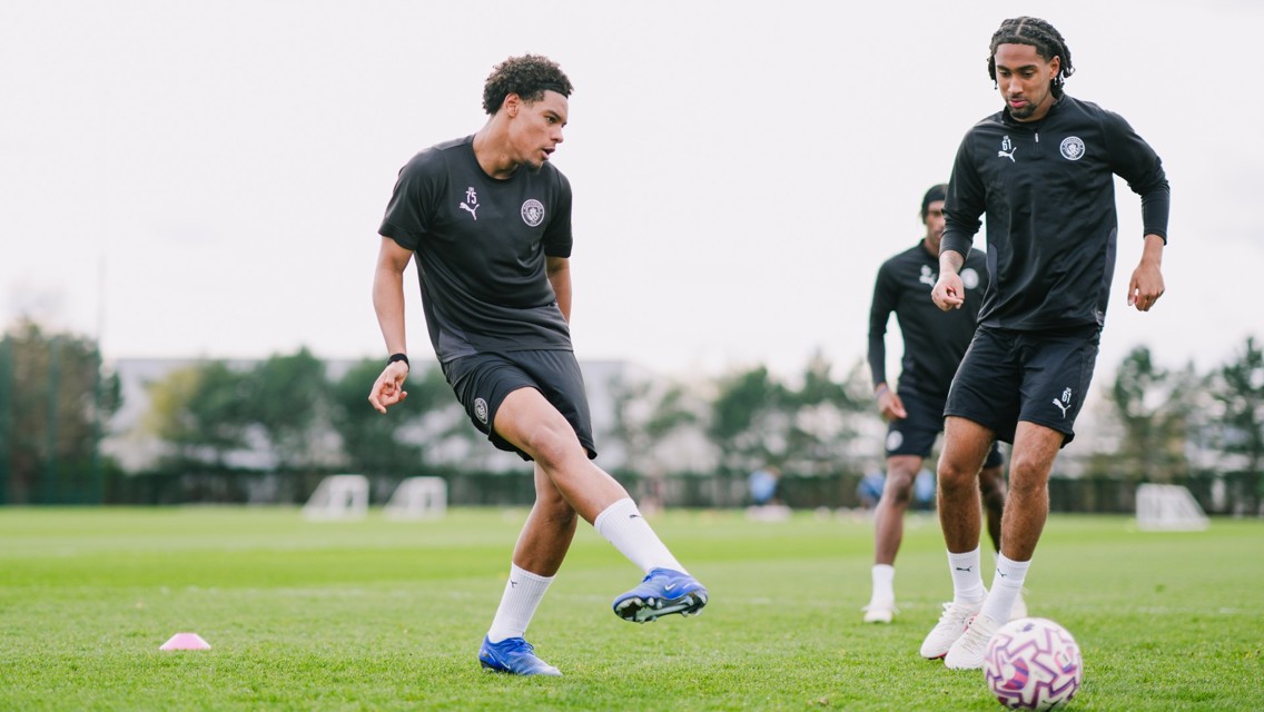 Three football players from Manchester City's youth team training on a grassy field, engaging in a dribbling exercise with a football.