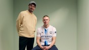 Two individuals, one standing in a sweater and cap, the other sitting wearing a Manchester City jersey in a green room.