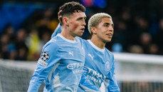 Two Manchester City players in light blue jerseys on the field, celebrating together with one placing a hand on the other's head, wearing UEFA Champions League patches.