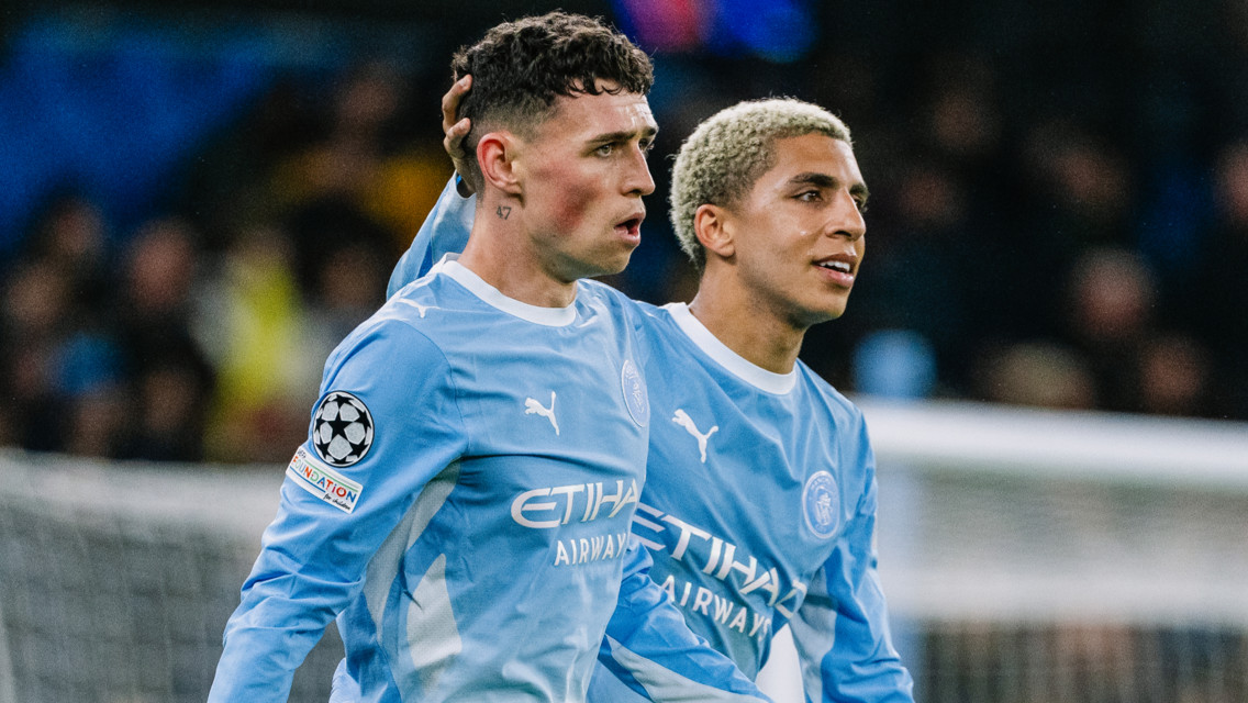 Two Manchester City players in light blue jerseys on the field, celebrating together with one placing a hand on the other's head, wearing UEFA Champions League patches.