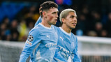 Two Manchester City players in light blue jerseys on the field, celebrating together with one placing a hand on the other's head, wearing UEFA Champions League patches.