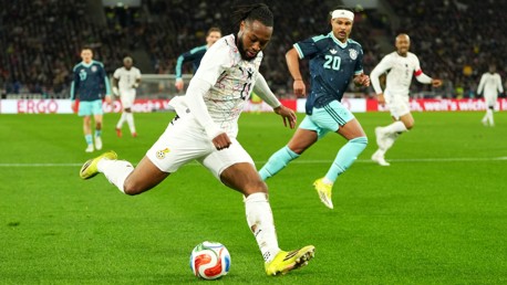 A soccer player from the team in white is about to strike the ball. He is closely followed by players from the team in blue. The match is taking place in a stadium filled with spectators.