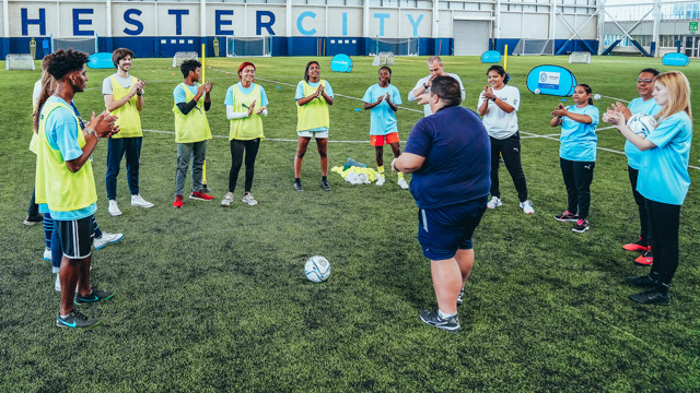 A group of young football players in blue and yellow bibs stand in a circle with a Manchester City staff member inside an indoor training facility. The signage 'HESTER CITY' is visible in the background.