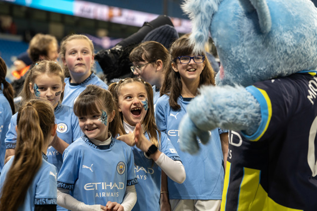 A group of young fans wearing Manchester City jerseys interact with the Manchester City mascot, Moonchester, on the field.