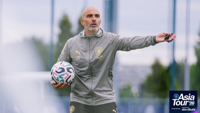A coach with 'EM' initials on his jacket holds a soccer ball while gesturing during Manchester City's Asia Tour 2026 in Abu Dhabi. The image includes the tour logo.