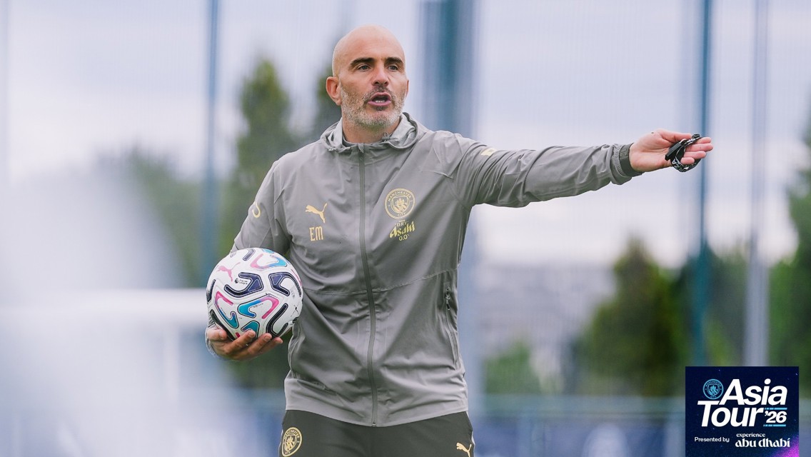 A coach with 'EM' initials on his jacket holds a soccer ball while gesturing during Manchester City's Asia Tour 2026 in Abu Dhabi. The image includes the tour logo.