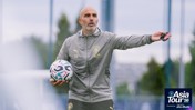 A coach with 'EM' initials on his jacket holds a soccer ball while gesturing during Manchester City's Asia Tour 2026 in Abu Dhabi. The image includes the tour logo.