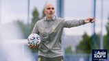 A coach with 'EM' initials on his jacket holds a soccer ball while gesturing during Manchester City's Asia Tour 2026 in Abu Dhabi. The image includes the tour logo.