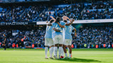 Manchester City players celebrate a goal at Etihad Stadium with a group hug. The stadium is full of fans watching the match.