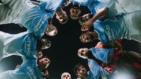 A group of football players in Manchester City uniforms huddle together, viewed from below with a focus on their team logos and sponsorships.