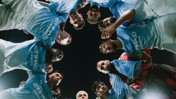 A group of football players in Manchester City uniforms huddle together, viewed from below with a focus on their team logos and sponsorships.