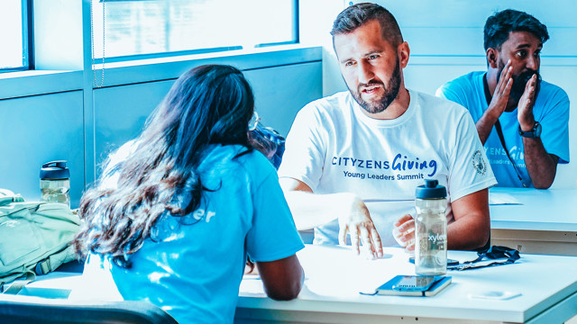 Two individuals at a table wearing 'CITYZENS Giving Young Leaders Summit' shirts engage in discussion in well-lit room. A backpack and water bottles are visible.