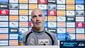 A man in a grey Puma jacket speaking at a press conference with a backdrop of sponsor logos. Two bottles are visible on the table, with the text 'Manager's Preview' and 'Asahi'.