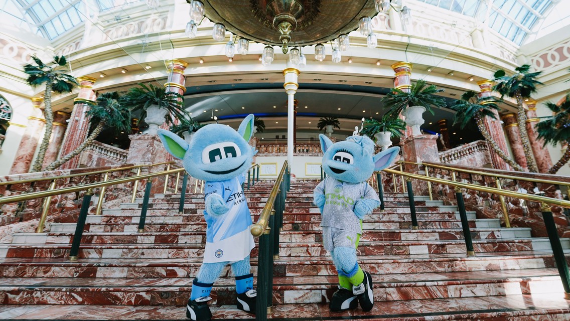 Two sky-blue mascots with large ears and monochrome eyes are standing on an ornate marble staircase with palm trees and a decorative ceiling in the background. They are wearing jerseys featuring Etihad Airways and Puma logos.