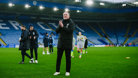 A football manager clapping on the field at King Power Stadium with others standing nearby, wearing winter coats. Players are visible walking in the background.