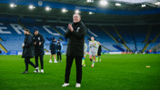 A football manager clapping on the field at King Power Stadium with others standing nearby, wearing winter coats. Players are visible walking in the background.