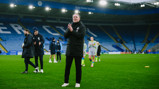 A football manager clapping on the field at King Power Stadium with others standing nearby, wearing winter coats. Players are visible walking in the background.