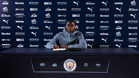 A person sitting at a table signing a document with Manchester City logos and sponsors in the background.