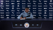 A person sitting at a table signing a document with Manchester City logos and sponsors in the background.