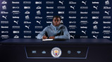 A person sitting at a table signing a document with Manchester City logos and sponsors in the background.