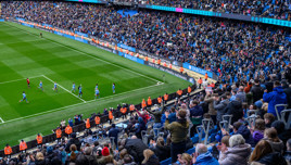 A large crowd of spectators in the stands at the Etihad Stadium watching a football match, with players in light blue uniforms celebrating a goal.