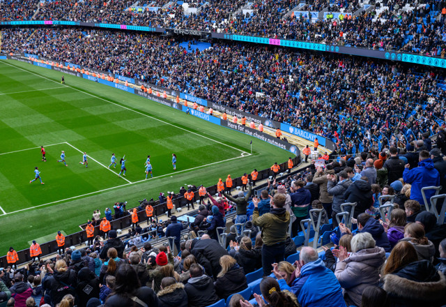 A large crowd of spectators in the stands at the Etihad Stadium watching a football match, with players in light blue uniforms celebrating a goal.