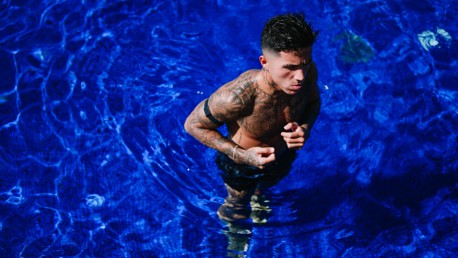A tattooed man with a serious expression is standing in a bright blue swimming pool, partially submerged in water.