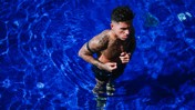 A tattooed man with a serious expression is standing in a bright blue swimming pool, partially submerged in water.