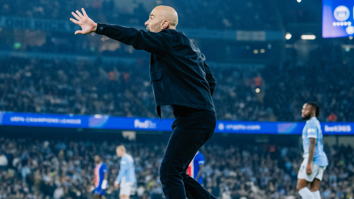 A bald man in a black jacket gestures passionately during a soccer match with players from Manchester City visible in the background playing in UEFA Champions League.